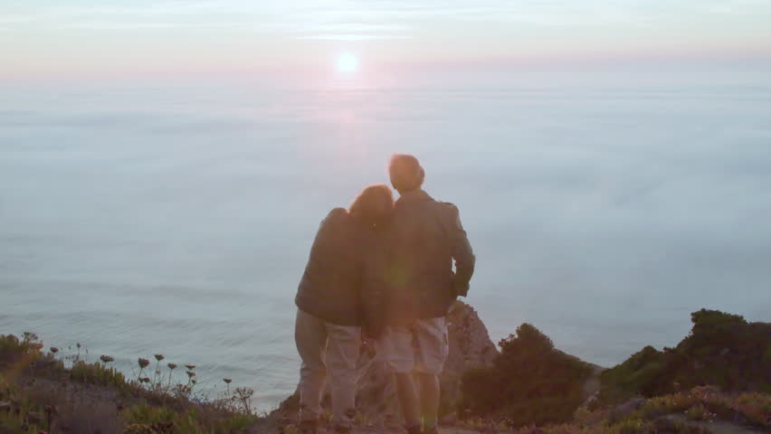 Senior couple standing on top of hill and admiring seascape. Back view of happy man and woman talking, holding hands, enjoying beautiful view after climbing mountain. Nature, love, hiking concept
