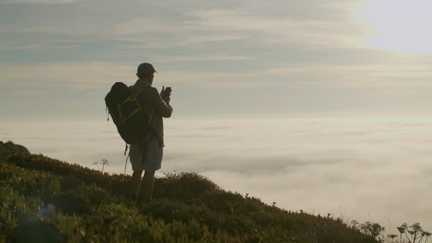 Hiker standing on mountain and taking photo of beautiful view. Long shot of senior man in cap enjoying landscape after climbing mountain at sunset, taking photo for social network. Hiking concept