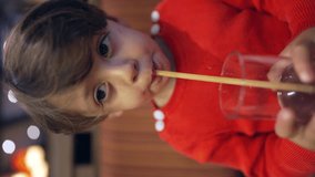 Child drinking from straw sitting at restaurant. Closeup child drinks beverage looking at camera. One thirsty little boy in Vertical Video