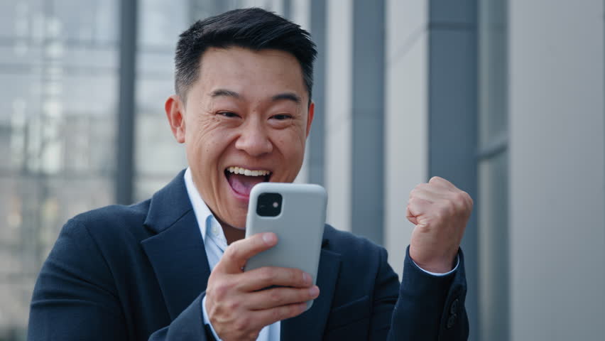 Close up Asian happy surprised man emotionally enjoys success good news 40s businessman boss win mobile online bet bid money financial prize victory receive offer opportunity on smart phone outdoors Royalty-Free Stock Footage #1098679429