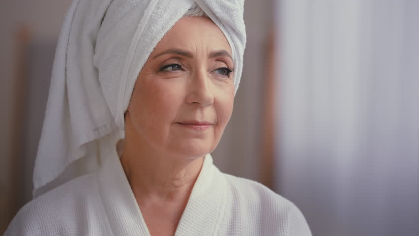 Close up female portrait middle-aged lady with towel on head in bathroom turn head to camera smiling 60s mature woman in bathrobe smile happy with facial skincare anti wrinkle cosmetics rejuvenation Royalty-Free Stock Footage #1098679443