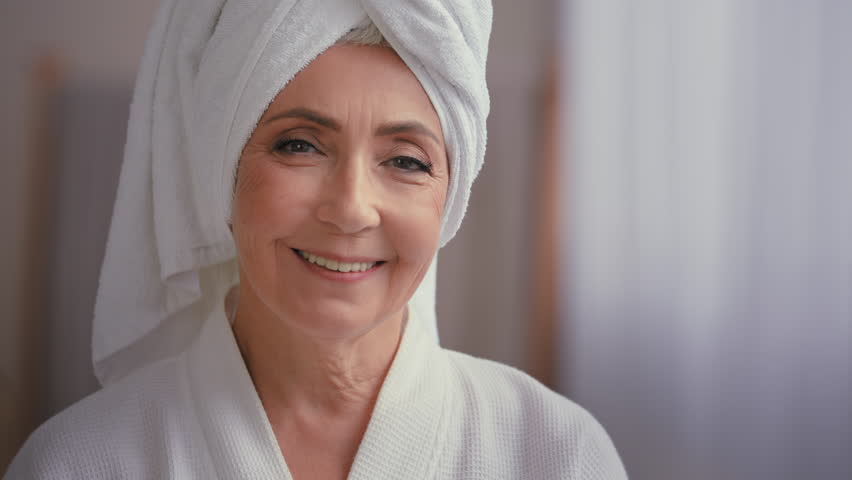 Close up female portrait middle-aged lady with towel on head in bathroom turn head to camera smiling 60s mature woman in bathrobe smile happy with facial skincare anti wrinkle cosmetics rejuvenation | Shutterstock HD Video #1098679443