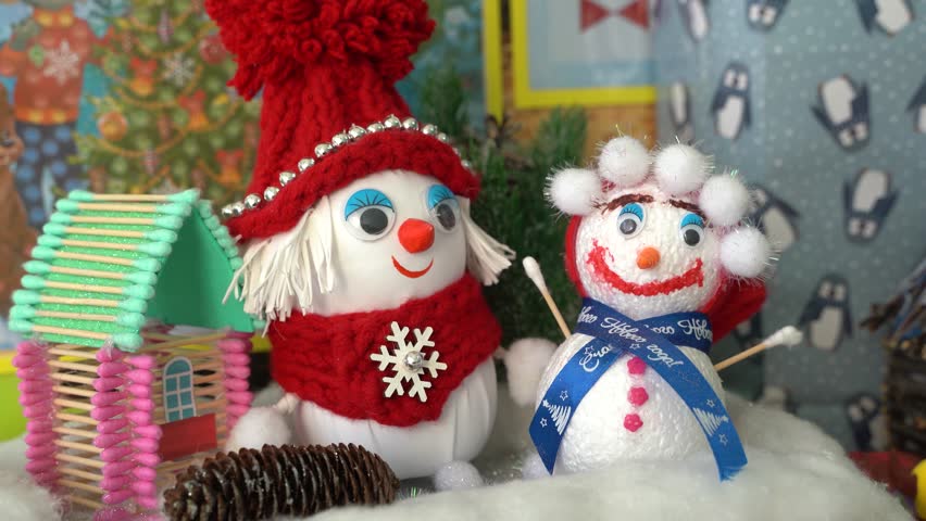 4K. Two soft toys-a boy and a girl snowman, stand on a cotton wool shelf for Christmas near a homemade green and pink house.