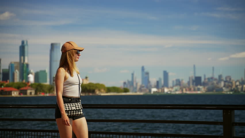 A video showing a woman walking and resting her hands on the black steel railing, looking at the New York city skyline across the river during daytime