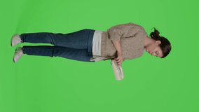 Vertical video: Side view of female model answering landline phone call on telephone line, standing over full body greenscreen. Happy girl using office phone, leisure activity on blank green screen