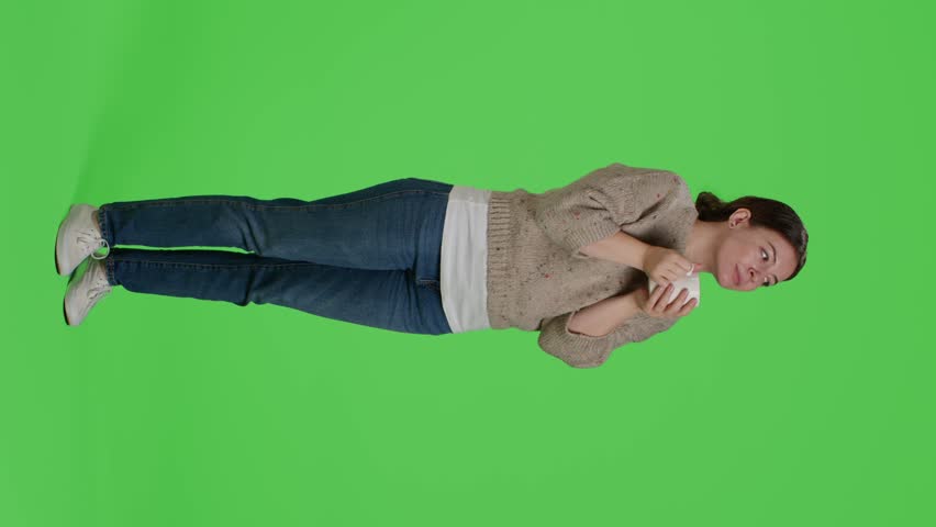 Vertical video: Side view of cheerful woman drinking cup of coffee in studio over full body greenscreen, holding hot drink. Young adult enjoying caffeine refreshment on isolated green screen backdrop.