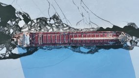 Aerial top down video of large bulk cargo ship moving through a frozen channel of water. Drone footage tracking above cargo ship traveling through icy waters.
