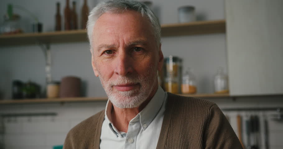 Bearded elderly man crosses arms turning to camera and smiling. Happy grey-haired pensioner in brown cardigan stands in kitchen at home closeup | Shutterstock HD Video #1098700469