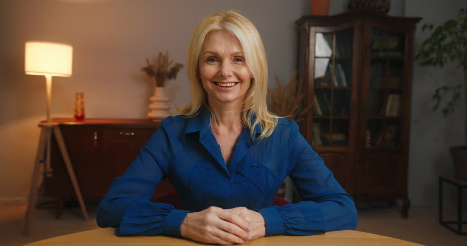 Mature blonde woman sits at table in office looking in camera and smiling. Successful female manager in blue shirt at business meeting closeup | Shutterstock HD Video #1098700501