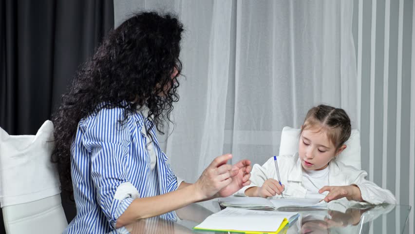 Nervous mother helps daughter with doing homework in apartment and explains material. Excited junior schoolgirl writes in notebook and listen to mom at table