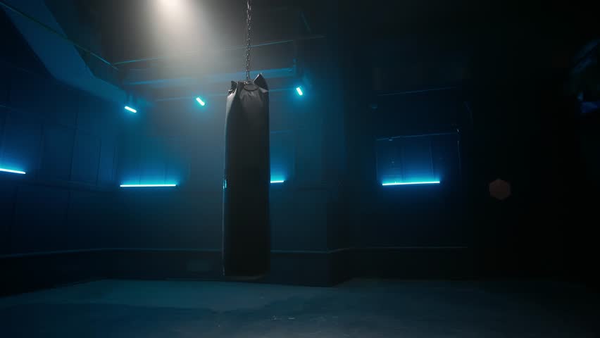 Long black punchbag hanging and swinging from chain in dark empty gym with blue neon lights. Leather punching bag for kickboxing, boxing, kicking and punching. Slow motion ready, 4K at 59.94fps. Royalty-Free Stock Footage #1098709877