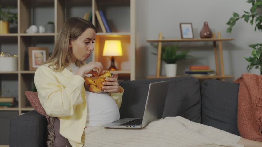 Beautiful expectant mother watching series on laptop and eating tasty snacks at home. Pregnant young woman relaxing on comfortable sofa with computer and fast food. | Shutterstock HD Video #1098713341