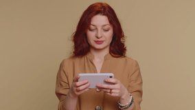 Worried funny addicted woman enthusiastically playing racing video games on mobile phone. Young redhead girl using smartphone gadget app with drive simulator isolated on beige studio background