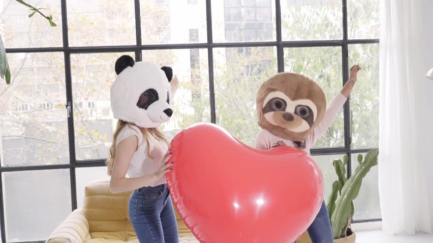 Two unrecognizable woman with animal mask bear panda holding big heart at home - Valentine