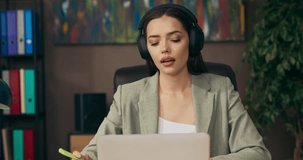 Serious buisness woman in suit wearing headphones sitting attable working from home discussing projects with colleagues online by video conference receiving call from director .