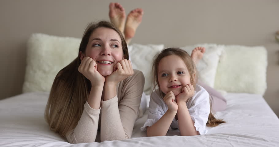 Carefree young mother her little cute daughter lying on bed, relaxing together in cozy bedroom, staring at camera, enjoy weekend look dreamy feel happy. Motherhood, childhood, family leisure at home Royalty-Free Stock Footage #1098725741