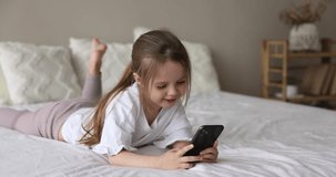 Cute little girl having fun on internet lying on bed using smart phone, watch videos, spend time playing on-line games. Young generation and modern wireless technology overuse, habit and lifestyle