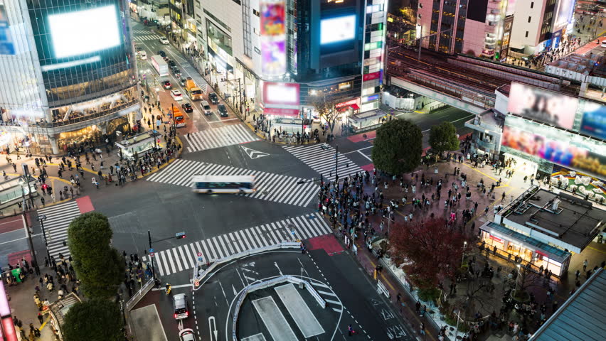 Night time lapse of car traffic transportation, crowded people walk cross road at Shibuya scramble crossing. All brands and logos blurred.