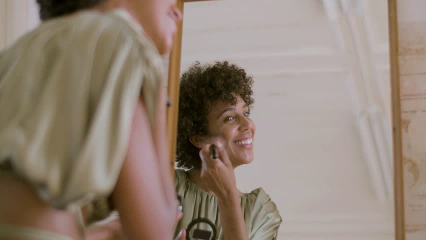 Happy Black woman doing make-up, applying powder on her face using make-up brush while sitting and smiling in front of mirror. Side view, low angle shot. Beauty, make-up concept.