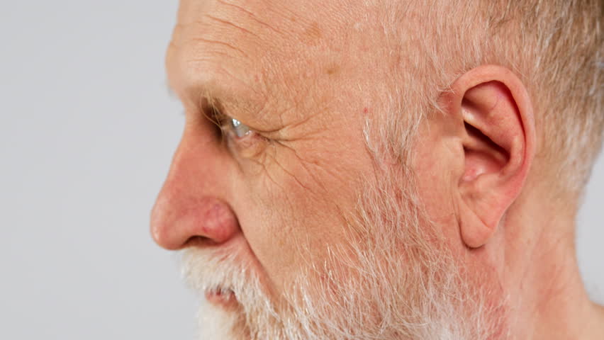 Close-up face of old man with long grey beard turns head and looking at camera.