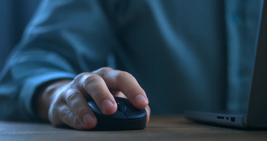 Close up man's hand uses a computer mouse while working on a laptop in the evening. Male freelancer clicking black mouse while browsing internet or working on computer remotely from home office. Royalty-Free Stock Footage #1098729357