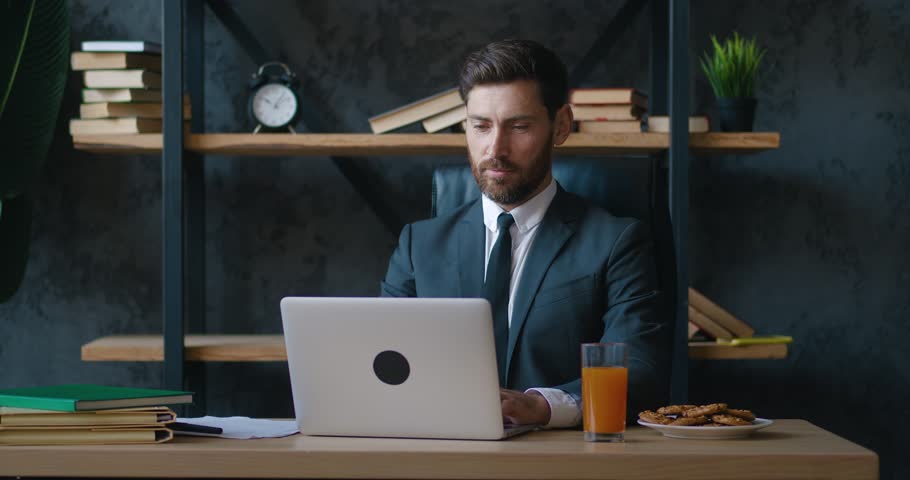 Confident bearded male businessman working on laptop and drinking juice in office. Professional male manager in a formal suit uses a computer while sitting at a table in a modern office | Shutterstock HD Video #1098729363
