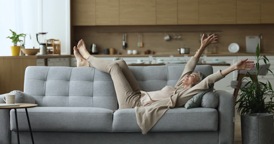 Happy carefree middle-aged female falling on cozy couch, smile enjoy comfort leisure at modern home with climate, control air-conditioning system. Stress-free weekend, relaxation, homeowner pastime | Shutterstock HD Video #1098732575