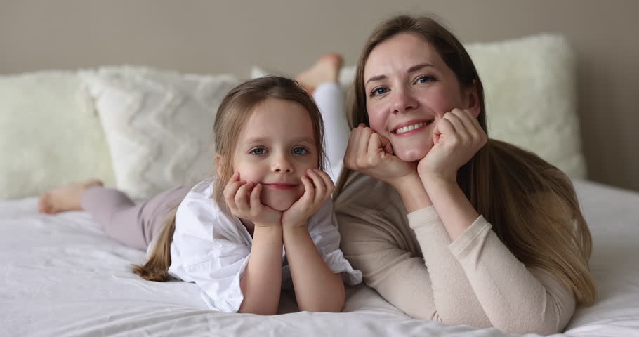 Happy mother and little daughter relaxing lying together on comfortable bed staring at camera, young woman spend lazy morning in cozy bedroom with awakened carefree smiling cute child at home. Family Royalty-Free Stock Footage #1098732599