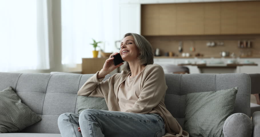 Attractive mature woman in comfortable casual clothes relax on cozy couch, making call to friend or children, talking on cellphone, smile enjoy pleasant personal conversation. Modern tech, phonecall Royalty-Free Stock Footage #1098732605