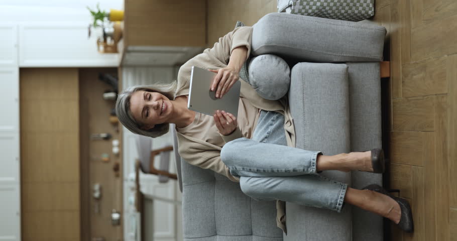 Smiling middle-aged woman resting with digital tablet, spend leisure alone sit on sofa in living room, buy goods, choose electronic services, enjoy easy comfort purchase on-line. Modern wireless tech | Shutterstock HD Video #1098732611