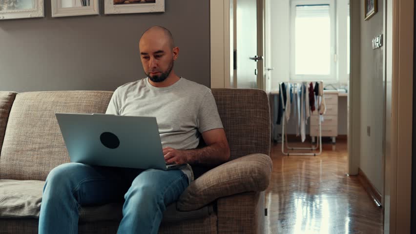 Caucasian bearded adult man walks and sits on sofa and working with laptop. Concept of freelance and remote work. | Shutterstock HD Video #1098736371