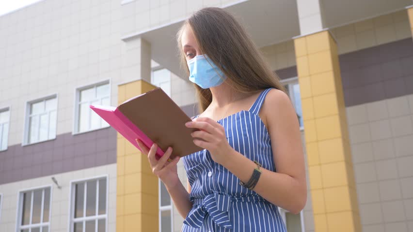 A girl reads a book in a Park with a medical mask on her face. A student is preparing for an exam remotely during the Covid-19 pandemic. a protective mask on the girl