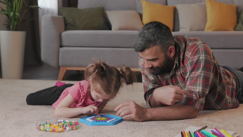 Young handsome father and cute daughter playing together. Dad and child making a puzzle with mosaic pegboard lying on the carpet at home. Educational toys for children. Home leisure. Father's day. Royalty-Free Stock Footage #1098747849