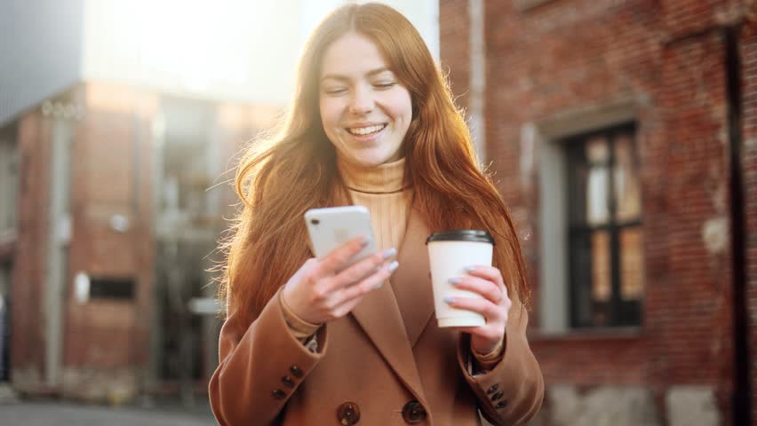 Portrait of smiling red haired ginger young woman with cup of coffee or tea hold smartphone scrolling social media texting browsing online on the street in brick city centre alone | Shutterstock HD Video #1098748653