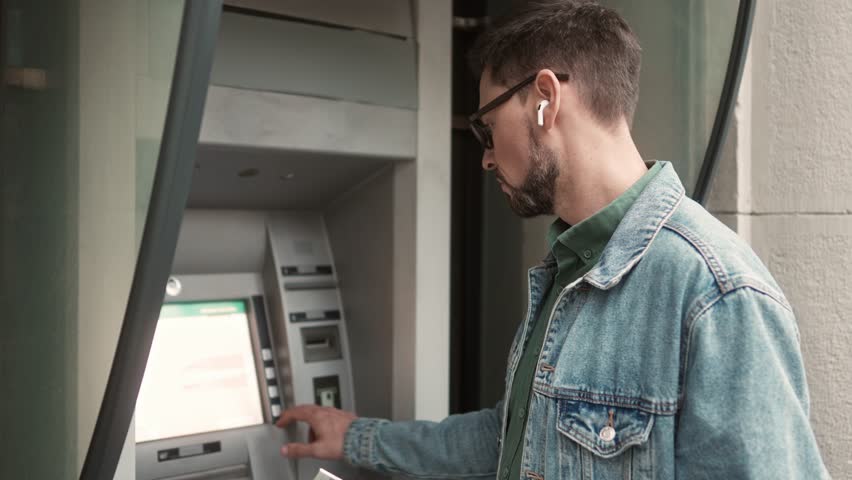Side-view portrait of young adult caucasian tourist using ATM to withdraw money from bank card check balance. Attractive modern man standing outside listening music in earphones holding map. Royalty-Free Stock Footage #1098749099