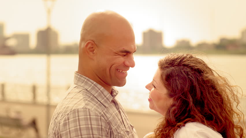 Attractive couple embracing by the Detroit River. Detroit and Windsor, Ontario, skylines visible in background.  Hand-held close up recorded at 60fps in 4K.  Clip tinted for additional golden warmth.