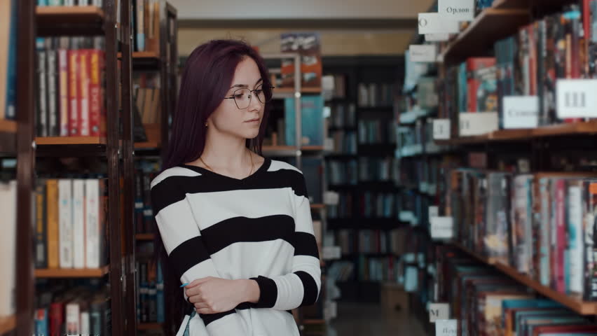 Caucasian female student with glasses looks at a shelf with books then into the camera, portrait. Female student in the library