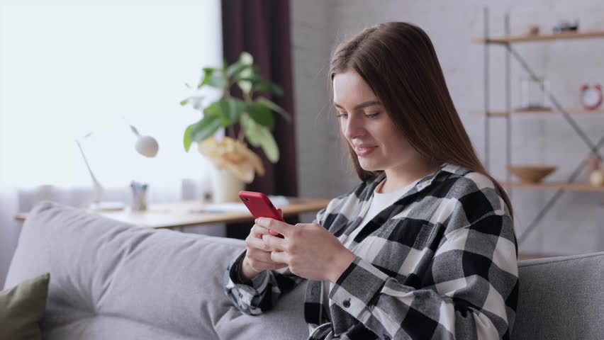 Portrait Beautiful girl tapping on sellphone at home. Relaxed female texting on smartphone, view photos and videos on the phone. Smiling young woman using smartphone in living room | Shutterstock HD Video #1098753791