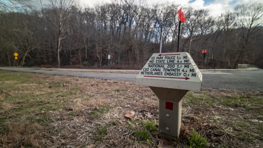 A time-lapse of a trail marker located near Beach Drive in Rock Creek Park in Washington DC. The camera rotates around the signpost, with motion-blurred people moving about in the background.