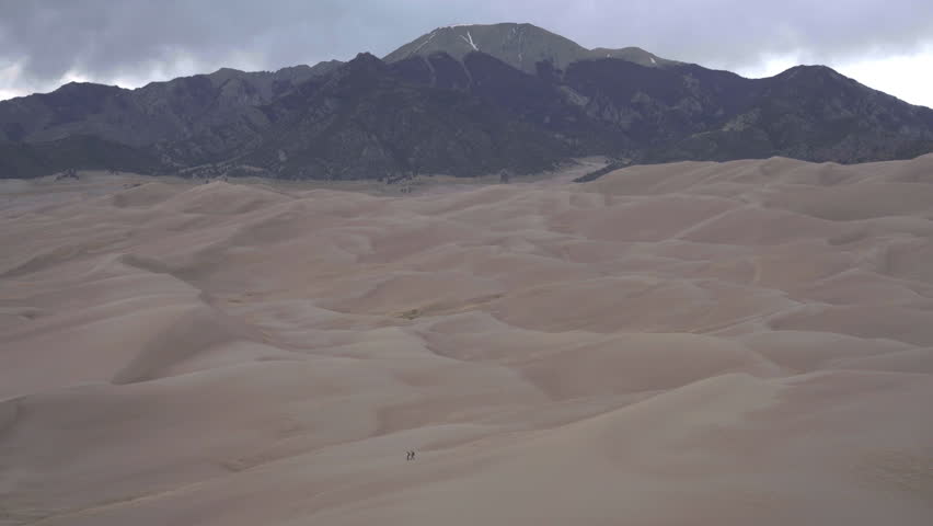 People walk across desolate sand dune field with mountains in back ground