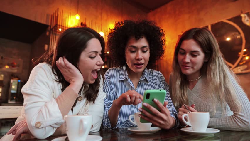 Group of young women having fun sharing media with an cellphone. Three girls looking to the smartphone on a coffee shop, restaurant or bar. Female students buying on a market place using a mobile Royalty-Free Stock Footage #1098768779