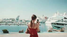 Cute woman takes photos, videos of the seaport on a mobile phone. girl uses social networks and streaming services on a smartphone when shooting yachts and ships