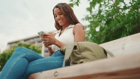 Beautiful girl with long dark hair wearing white top sits on bench and uses mobile phone. Young smiling woman writing in social networks on cellphone. Sideways movement