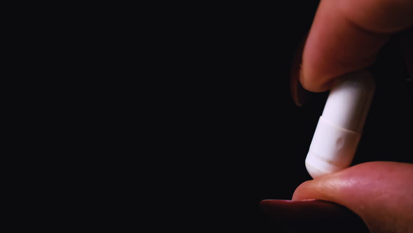 Woman holds a capsule pill in her hands close-up on a black background. The girl examines the mountain of capsules that she has in her hand. A woman stands in her house, apartment with medicine in her | Shutterstock HD Video #1098771205