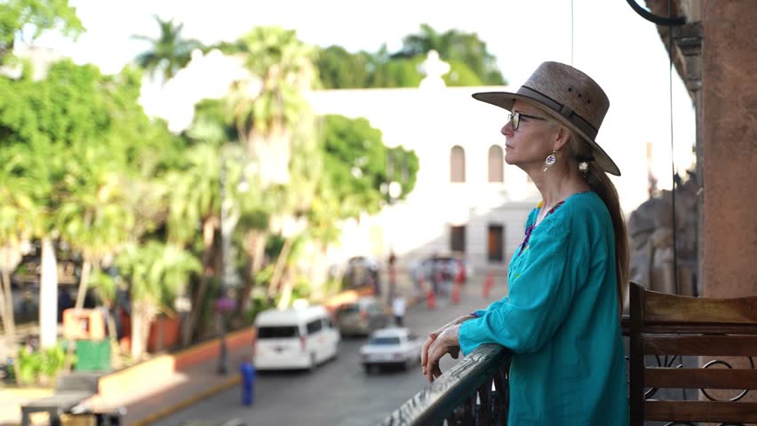 Closeup of smiling happy mature woman wearing a hat, looking out from a balcony over the park in Merida, Yucatan, Mexico.