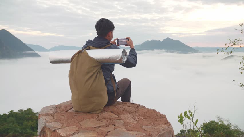 Asian Hiker Male Sitting On The Rock And Using Smartphone Taking Photo Of The Top View Foggy Mountain
