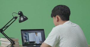 Close Up Back View Of Young Asian Male Raising His Hand To Ask Question While Studying On Green Screen Background And Looking At A Male Teacher Teaching On A Laptop At Home
