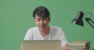 Close Up Of Young Asian Male Waving Hand For Greeting And Speaking While Studying On A Laptop On Green Screen Background At Home
