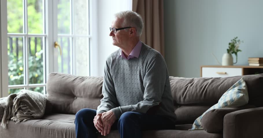 Sad unhappy elderly retired man sits on couch alone, sighs, looks out window feels abandoned, suffer from weakness, ageing disorders, grieving about past, missing for grown up children living far away | Shutterstock HD Video #1098773607
