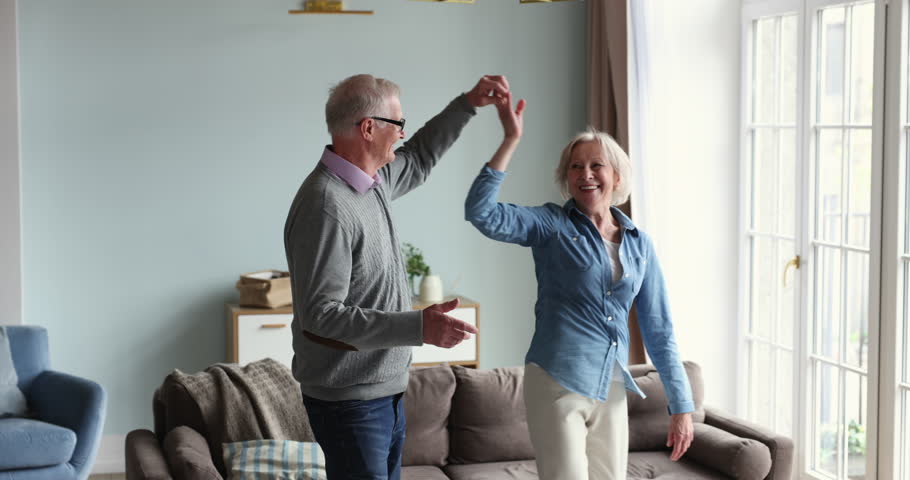 Active older couple spend carefree leisure together at home, listen favourite song, holding hands dancing, feel happy. Retired couple in love enjoy romantic date move to music have fun in living room Royalty-Free Stock Footage #1098773651
